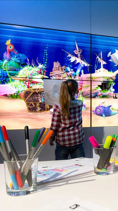 Ein Mädchen steht vor dem paint2life Aquarium und beobachtet ihren gemalten Fisch