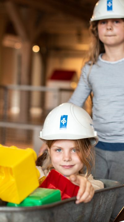 Mädchen mit Bauhelm schiebt anderes Mädchen in Schubkarre