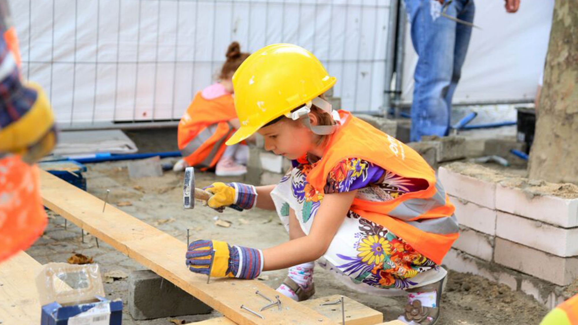 XXL Baustelle Kind mit Bauweste und Bauhelm schlägt Nagel in Brett